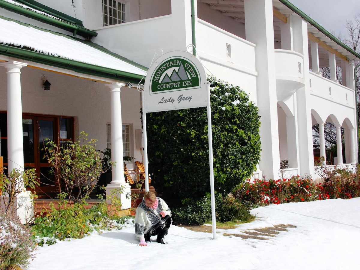 An inviting country inn is depicted, featuring a prominent sign that identifies the establishment. Snow blankets the ground, enhancing the serene atmosphere. Lush greenery and vibrant flowers border the entrance, while the building's elegant architecture is partially visible, showcasing tall columns and large windows.