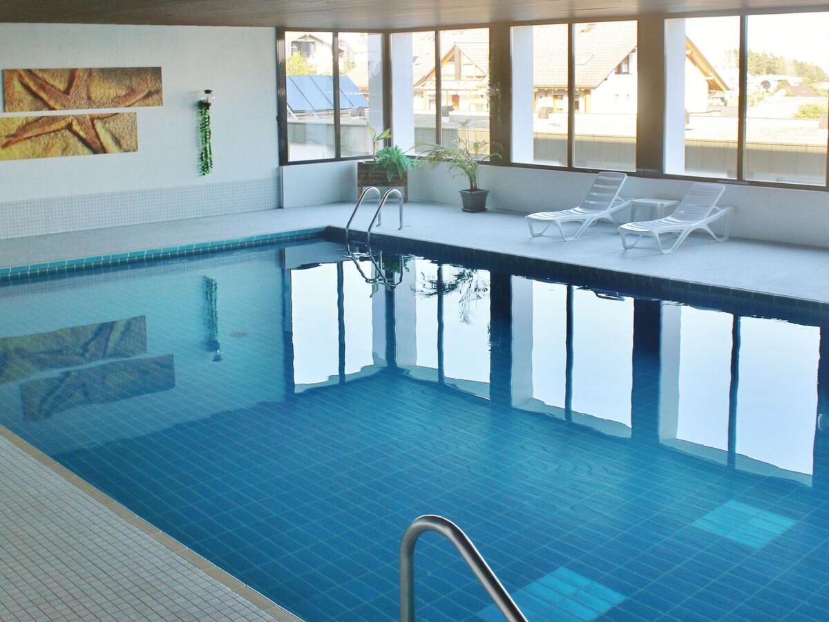 An indoor pool is featured, showcasing clear blue water reflecting the light from the surrounding windows. Two lounge chairs are positioned nearby, with a potted plant adding a touch of greenery. The area is well-lit, creating a calm and spacious environment.