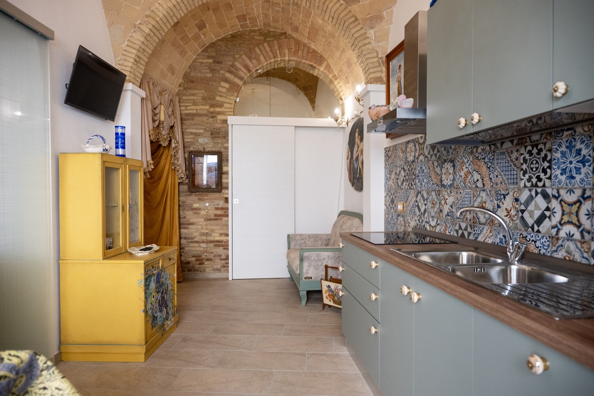An open-concept living space features a kitchen area with a dual-basin sink and contemporary cabinetry. Textured walls display traditional tiles, while an arched ceiling adds historical character. A seating area is provided, complemented by a cabinet that offers storage and decorative elements.