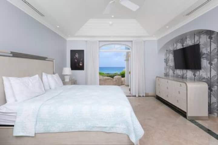 غرفة نوم رئيسية جميلة مع سرير أمريكي بحجم كينج ومكيف هواء ومروحة سقف وتلفزيون وأبواب فرنسية تؤدي إلى الحديقة وحمام داخلي مجهز جيدًا.