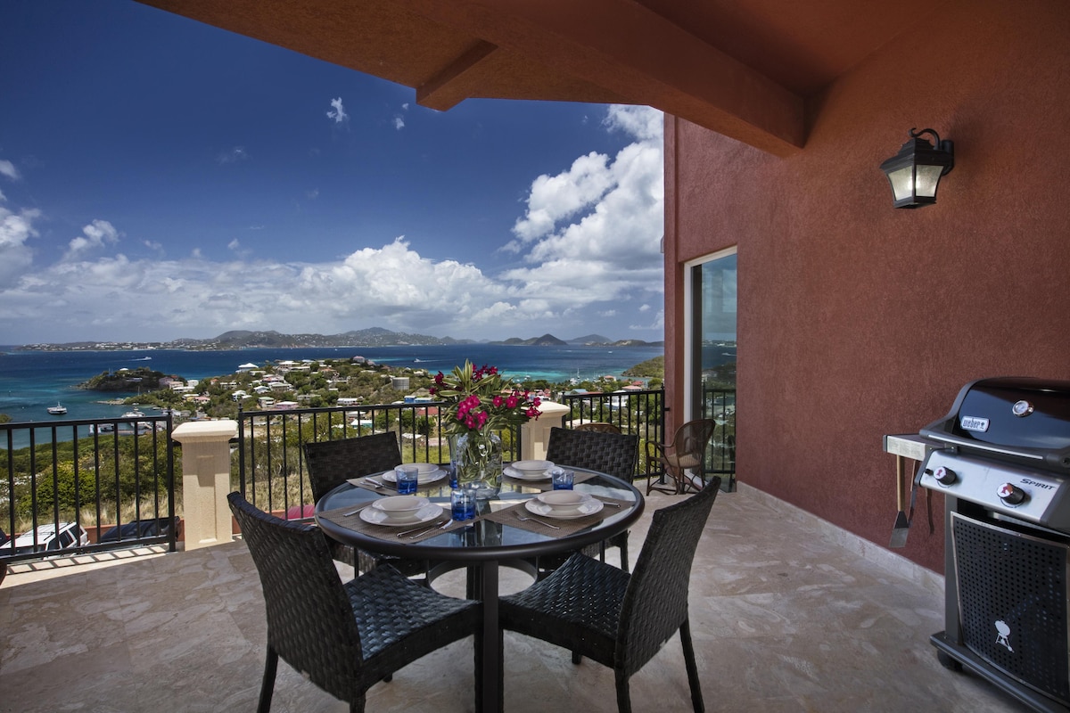 An outdoor dining area is observed with a round glass table and four woven chairs, set against a vibrant backdrop of sweeping ocean views. A gas grill is positioned nearby, while colorful flowers decorate the table, enhancing the inviting ambiance of the patio.