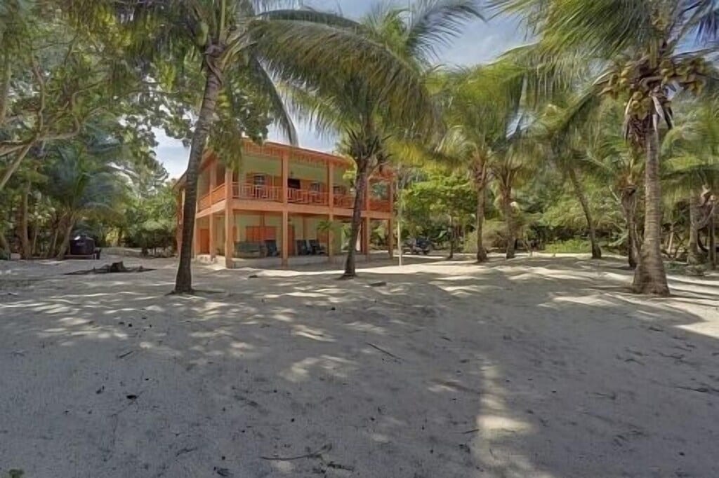 A two-story beach house is nestled among swaying palm trees, surrounded by sandy ground. The structure features a welcoming orange exterior and a wraparound veranda, providing a relaxing space to enjoy the outdoor environment. Sunlight filters through the leaves, creating dappled shadows on the beach.