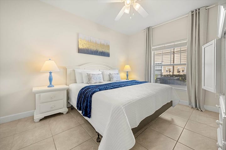 Este dormitorio luminoso y espacioso cuenta con una cómoda cama tamaño king con sábanas blancas y una acogedora colcha azul marino. La iluminación suave junto a la cama agrega calidez y las grandes ventanas dejan entrar la luz natural al tiempo que ofrecen hermosas vistas del exterior.