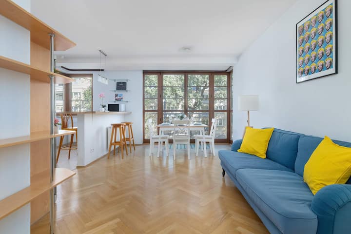 Svetlá obývačka s modrou pohovkou a jedálenským kútom viditeľným v pozadí. Vďaka veľkým oknám je priestor vzdušný a priestranný.