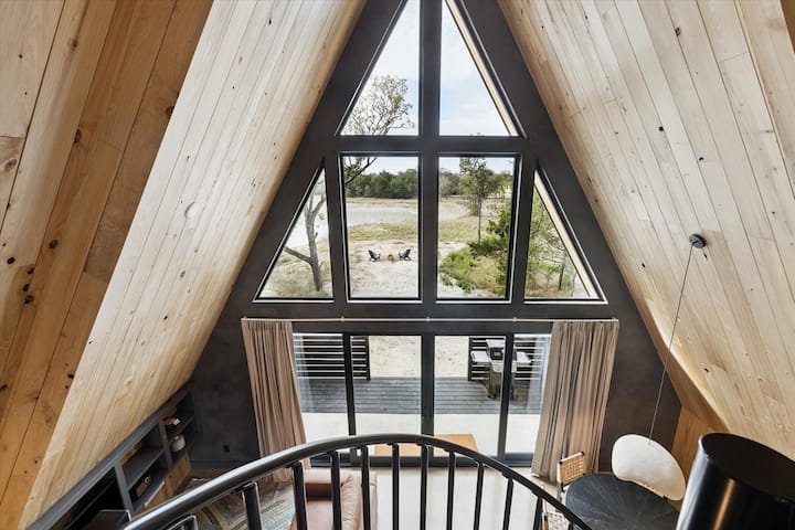 Спалната соба на поткровјето сместена во високите прозорци на таванот од подот до таванот на A - рамката го врамува погледот кон шумата, додека изложените греди и меката постелнина создаваат удобно, издигнато место за одмор.