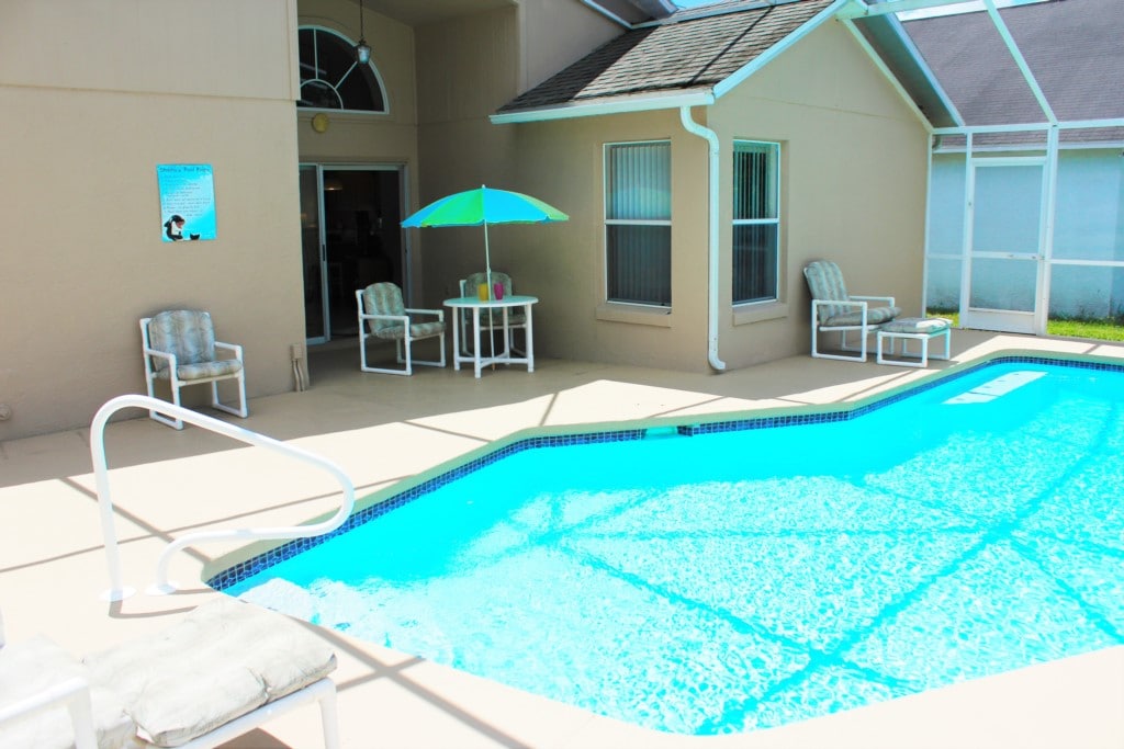 This outdoor area features a private pool surrounded by a spacious deck. Comfortable lounge chairs are positioned nearby, along with a table and an umbrella providing shade. Clear windows from the adjacent room offer a view of the pool area.