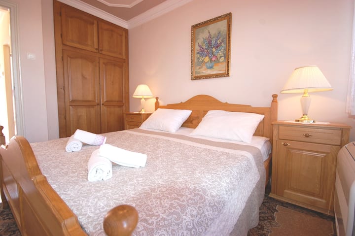 Un acogedor dormitorio con una cama de madera, ropa de cama y toallas cuidadosamente dispuestas, mesitas de noche con lámparas y un gran armario. El espacio se ve cálido, ordenado y acogedor.