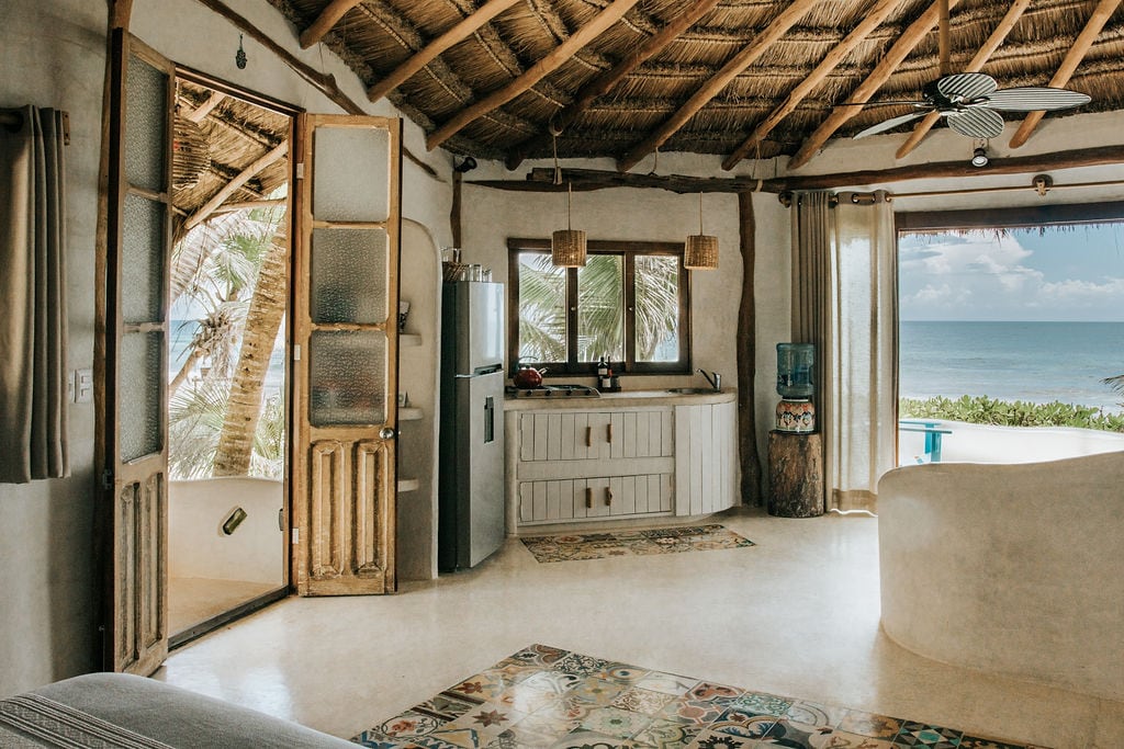 An open-concept living space features a combination of wooden doors and windows that frame views of the turquoise sea. The kitchen area includes a fridge and a sink, while soft natural light illuminates curved walls and a textured ceiling of exposed beams.