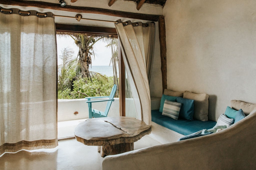 An inviting living area features a curved wall and natural materials, with a wooden table at the center. Eco-friendly furniture includes a comfortable seating area with cushions and an accent chair positioned to offer views of the sea through sheer curtains that softly diffuse the light.