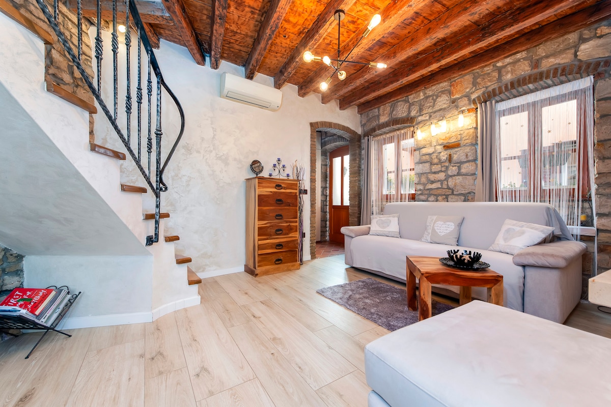 A living room features a comfortable sofabed and a wooden coffee table, set against a backdrop of exposed stone and warm wooden beams. Natural light filters through the tall windows, enhancing the welcoming ambiance. A staircase leads to the upper level, adding architectural interest.