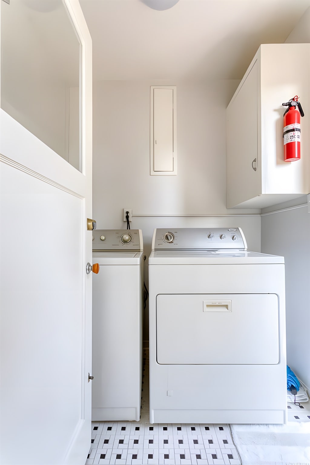 A laundry room features a stacked washer and dryer in white. Adjacent to them, storage cabinets provide additional space, and a fire extinguisher is mounted on the wall. The floor is accented with small black and white tiles, creating a clean, functional space.
