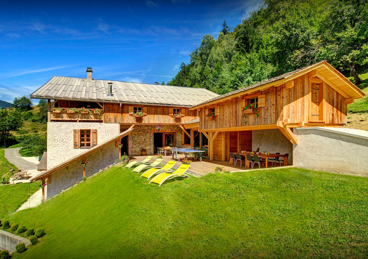 The exterior of the chalet features rustic wooden architecture set against a lush green hillside. A spacious terrace is adorned with sun loungers and dining tables, surrounded by landscaped gardens. The design highlights natural materials and integrates harmoniously with the surrounding landscape.
