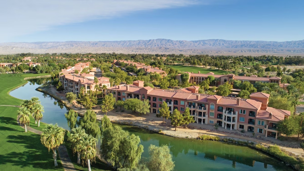 The resort is surrounded by expansive green lawns and palm trees, alongside a serene water feature. Mountain ranges can be seen in the background under a clear blue sky, adding to the tranquil setting of the Palm Desert landscape.