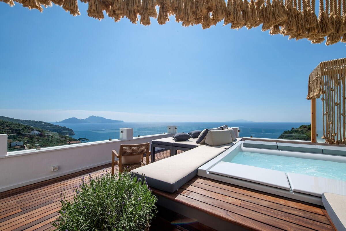 A sunlit terrace features a stylish lounge area with sun beds and a mini-pool. The wooden flooring complements the view of the sea and distant islands. Decorative elements include woven accents, enhancing the serene outdoor space.