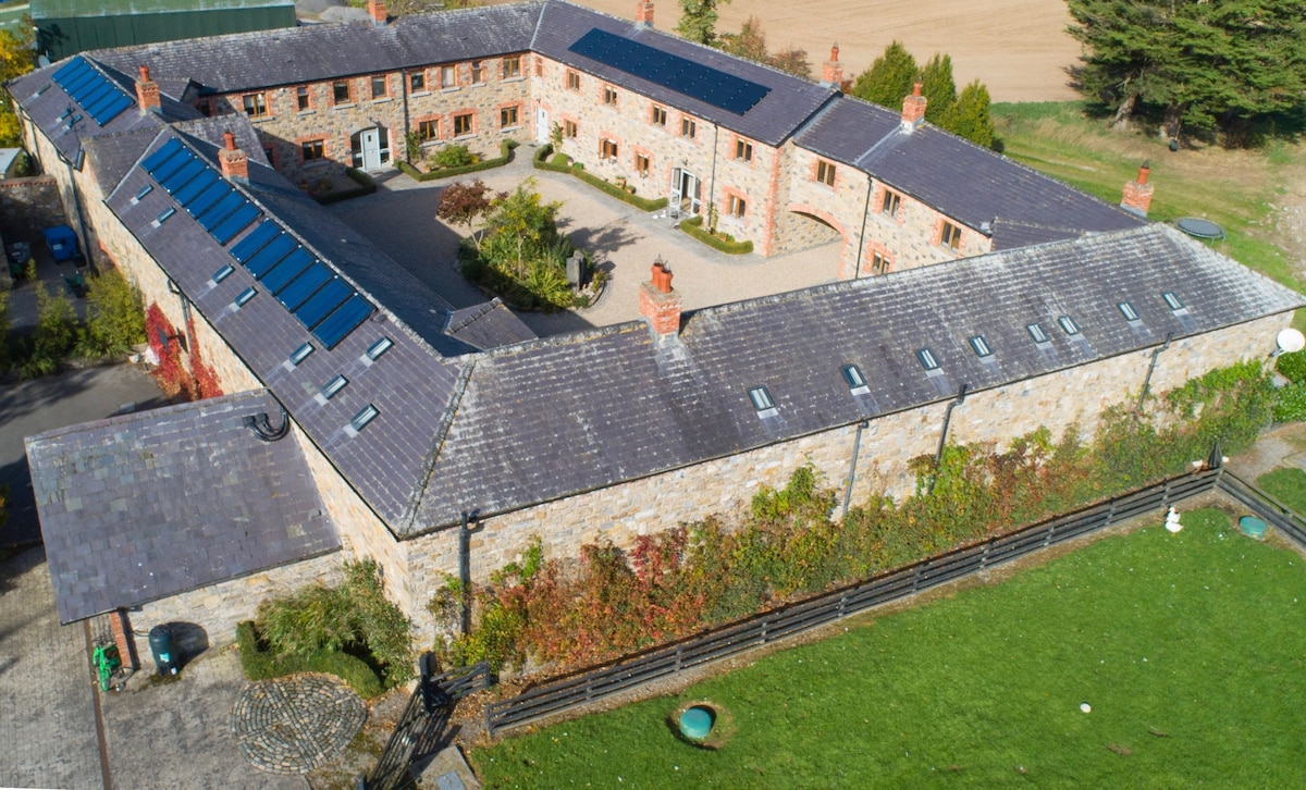 An aerial view of the landscaped courtyard reveals a charming stone building with a slate roof, featuring multiple windows and solar panels. The surrounding grounds include a grassy area and trees, creating a serene and inviting environment.