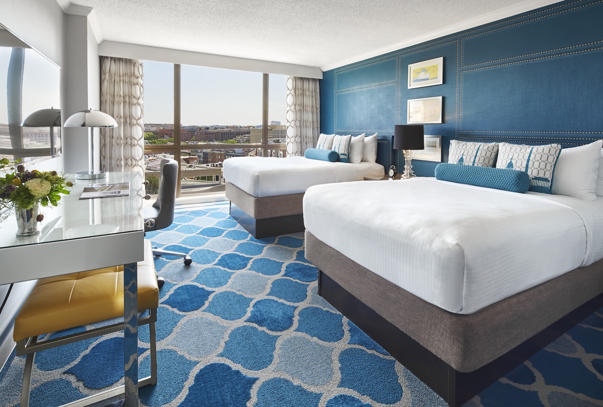 Двойни легла с яркосиня стена, работно бюро и прозорци с изглед към града