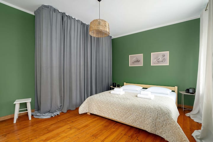 منظر آخر لغرفة النوم مع السرير المزدوج، يُظهر الستائر ومقعد صغير بجانب السرير. المسكن يبدو هادئًا ومشرقًا.