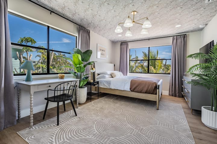 Stijlvolle slaapkamer met grote ramen, weelderig uitzicht en natuurlijk licht. Beschikt over een knus bed, een moderne inrichting en een chique werkruimte, perfect om te ontspannen of om je werk bij te werken in een serene, op de kust geïnspireerde omgeving.