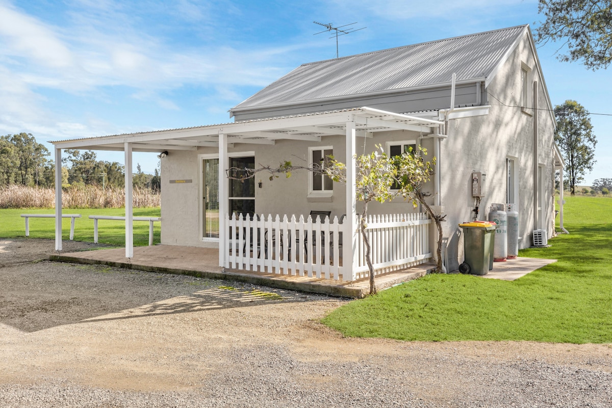 A charming, single-story cottage features a white picket fence and a spacious porch area. The exterior is lightly colored, complemented by a metal roof. Lush green grass surrounds the property, with a gravel path leading up to the entrance. Nearby, waste bins and a water source are visible.