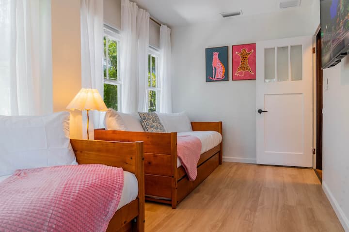 Светла спална соба со два кревета и сончеви прозорци, разиграна уметност и пријатна минималистичка удобност.