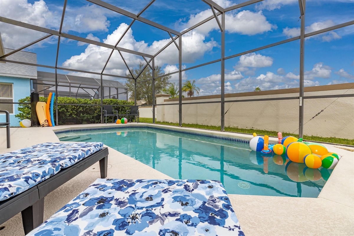 A private pool area is enclosed with a screened lanai, featuring a clean, inviting pool surrounded by colorful pool toys. Comfortable loungers with floral cushions are positioned near the water, and a clear blue sky with fluffy white clouds is visible above.
