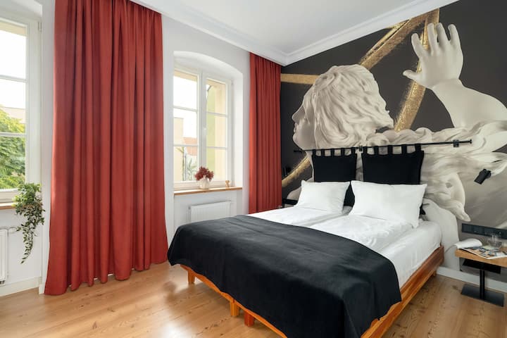 Chumba maridadi cha watu wawili kilicho na kitanda kizuri, matandiko ya kifahari meusi na meupe na mapazia mekundu yenye ujasiri. Ukuta wa kisanii ulio na sanamu za zamani unaongeza sifa ya kipekee kwenye sehemu hiyo.