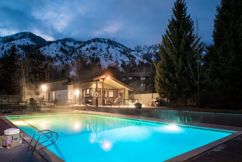 A heated outdoor pool is set against a backdrop of snow-capped mountains. Soft lighting from nearby structures reflects off the water, creating a serene ambiance. Lounge chairs surround the pool, and wisps of steam rise, indicating the warmth of the water.