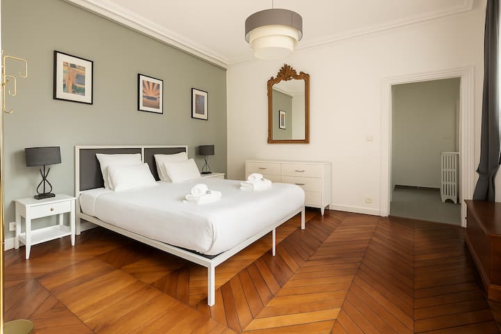 
Habitación espaciosa y luminosa de estilo depurado. La pared verde salvia, los muebles blancos y el suelo de parqué en espiga crean un ambiente elegante y relajante. La cama doble, las lámparas negras y el espejo dorado aportan comodidad y refinamiento.