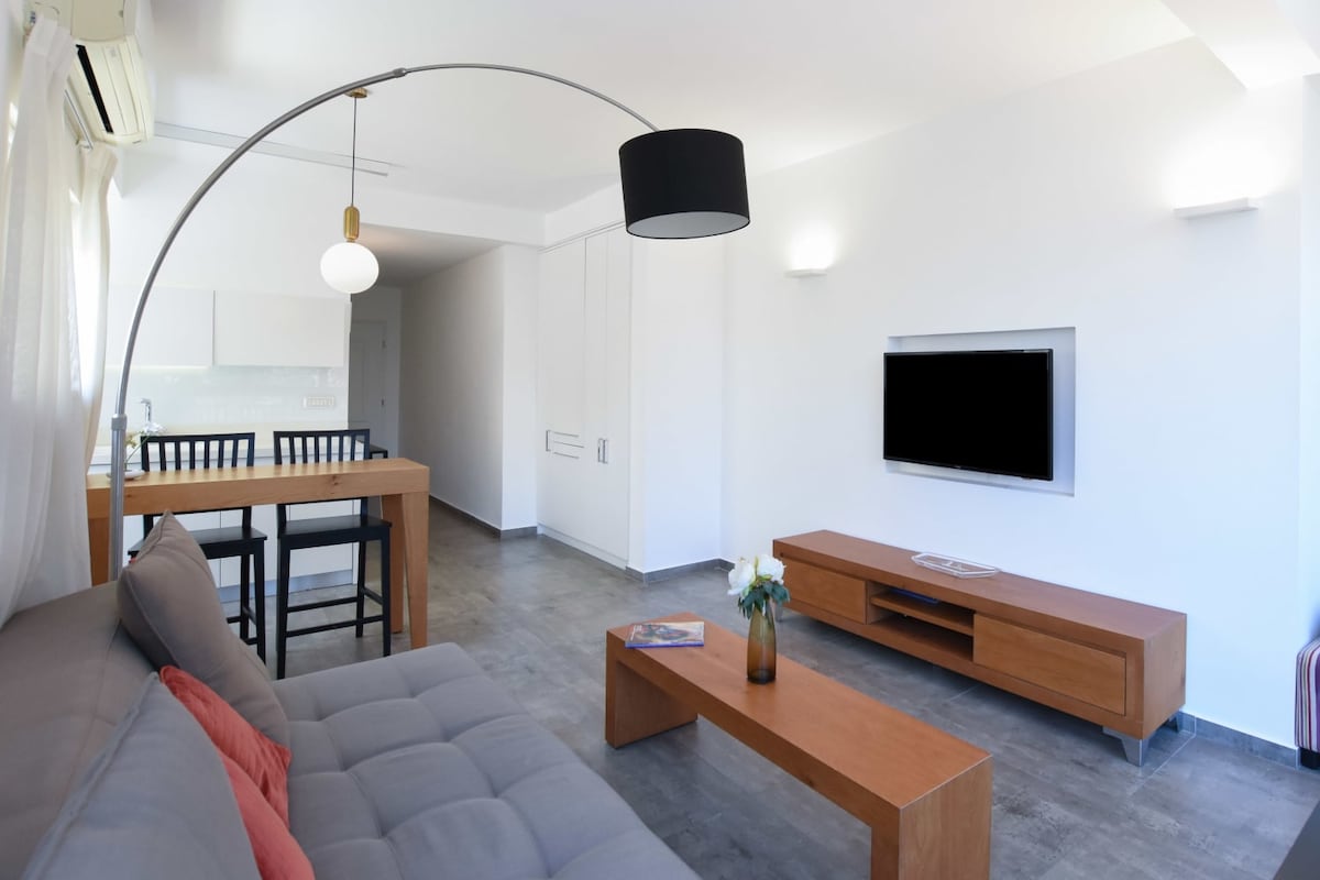 A spacious living area features a grey sofa with decorative pillows, complemented by a wooden coffee table. A TV is mounted on the wall across from the seating, with a stylish arch lamp providing ambient lighting. The adjacent kitchen area is partially visible, showcasing a dining table and chairs.