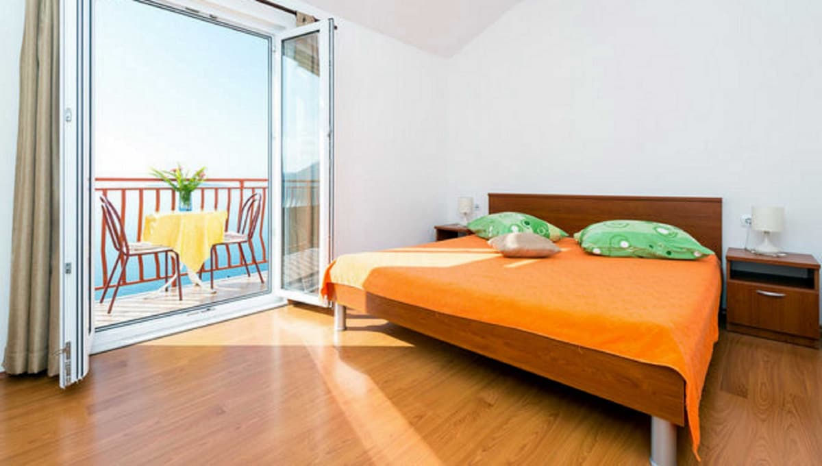 A bright bedroom features a spacious bed topped with a vibrant orange coverlet and decorative pillows. Double glass doors open to a balcony with a small table and chairs, offering views of the sea. Natural light fills the room, highlighting the wooden flooring.