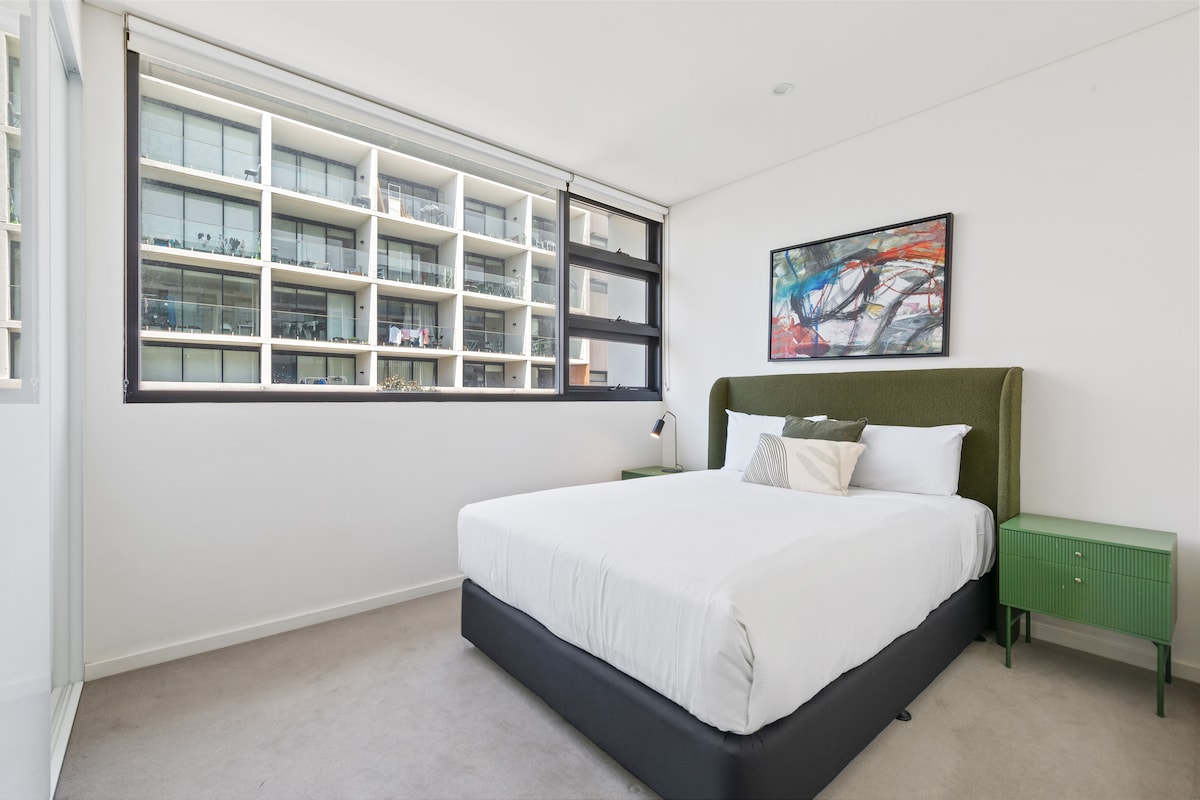 Een rustgevende slaapkamer met queensize bed die de strakke esthetiek van het appartement weerspiegelt, met pluche beddengoed, moderne verlichting en grote ramen die de ruimte in natuurlijk licht baden.