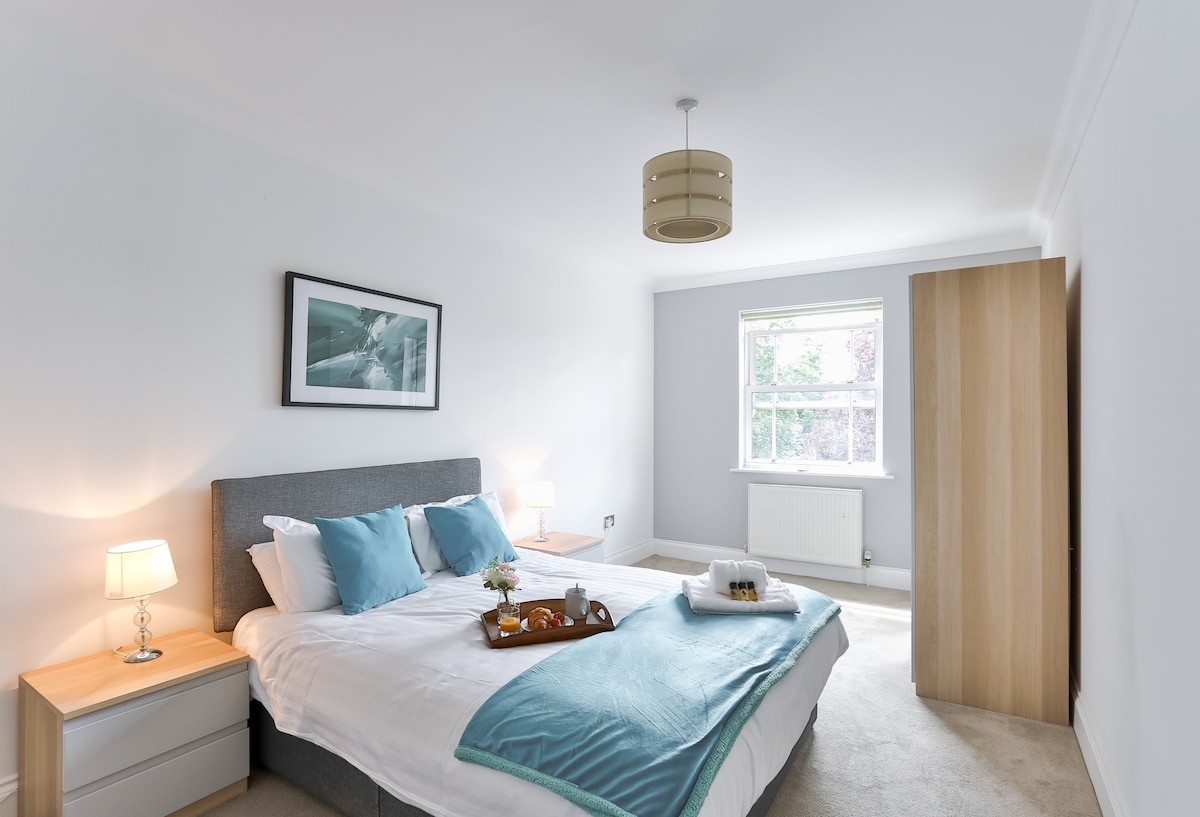 A spacious bedroom is presented with a comfortable bed featuring soft pillows and a throw blanket. Bedside tables with lamps frame the bed, while a window allows natural light to brighten the neutral-toned walls. A wardrobe stands to the side, enhancing the room's functionality.
