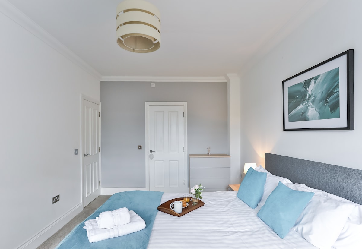 A bright bedroom features a spacious bed adorned with white linens and plush turquoise accents. An elegant wooden tray holds a small vase with flowers, positioned atop the bed. Soft light streams in through the window, complementing the calming grey walls.