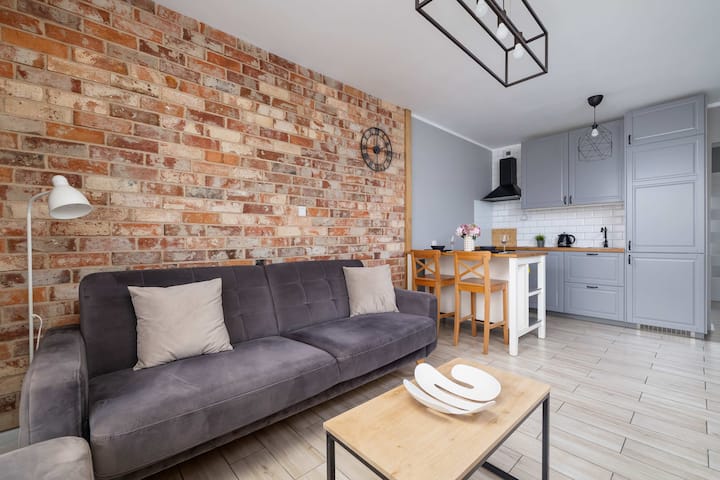 Un salon confortable avec un canapé gris et une petite table basse, avec des murs en briques apparentes pour une ambiance chaleureuse. La cuisinette est visible en arrière-plan.