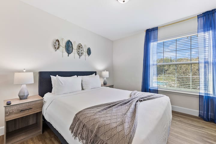 მსუბუქი საძინებელი, რომელშიც არის საწოლი 152×203 სმ ზომის, ლურჯი ელემენტები, დეკორატიული ნამუშევრები და კომფორტული თეთრეული.