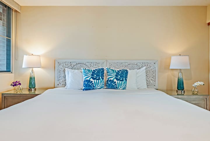 Une chambre avec une tête de lit à motifs et des oreillers bleus, la lueur des lampes créant une ambiance chaleureuse et calme tandis que les vagues chuchotent à l'extérieur.
