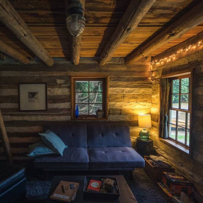 The cabin interior features exposed wooden beams and rustic log walls. A dark loveseat is positioned near two windows, providing a view of the outdoors. A small table holds a few items, while a lamp and string lights add subtle illumination to the space.