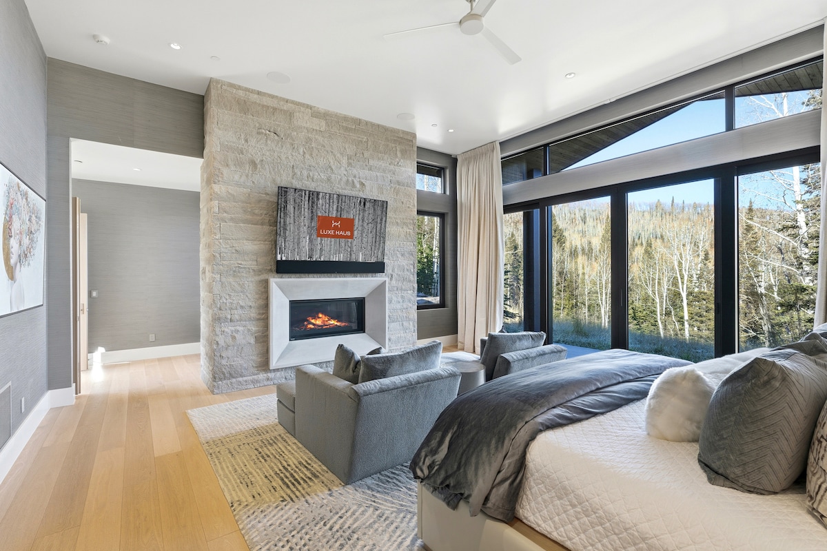 Retiro de dormitorio en esquina con chimenea de piedra, rincón de lectura y ventanas panorámicas que ofrecen vistas a las montañas boscosas.