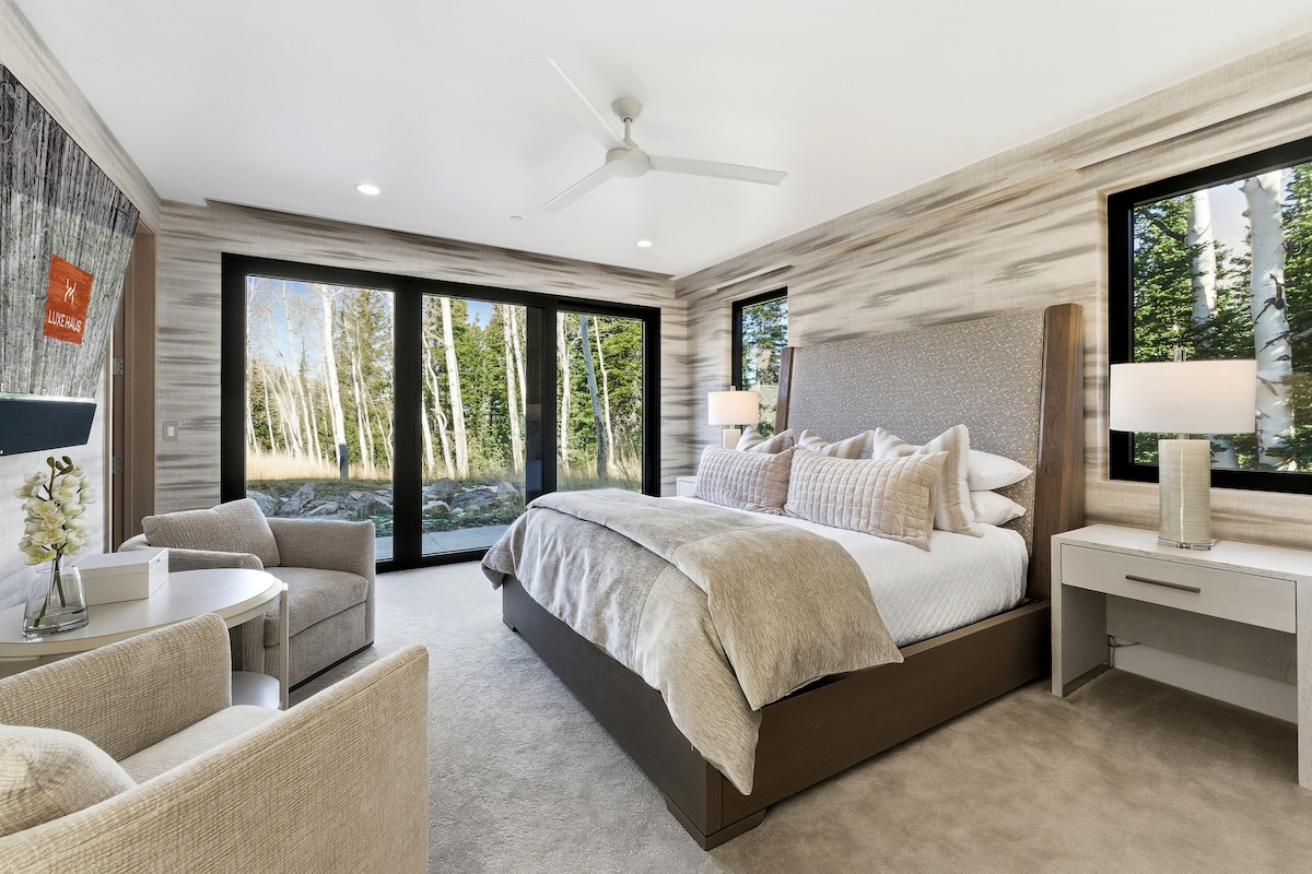 Suite de invitados luminosa con grandes ventanales, cama tapizada y zona de estar con vistas al bosque.