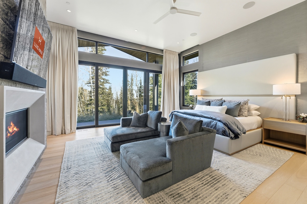 Amplio dormitorio con asientos de salón, vistas a la montaña y grandes ventanales que enmarcan el entorno alpino.