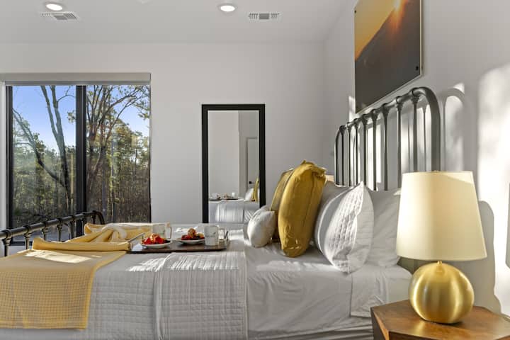 This bright master bedroom features a stunning forest view, a sleek metal bed frame, and warm gold accents throughout.
