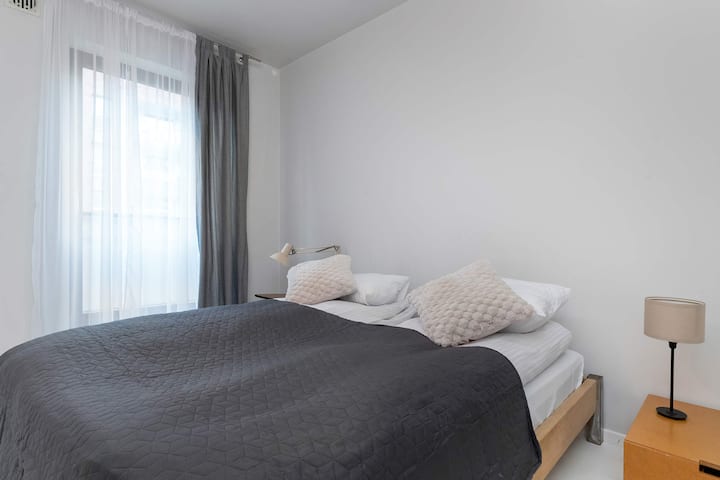 Un pequeño dormitorio con una cama doble cubierta con ropa de cama gris suave. Las cortinas transparentes difunden la luz natural. El espacio se ve tranquilo y sencillo.