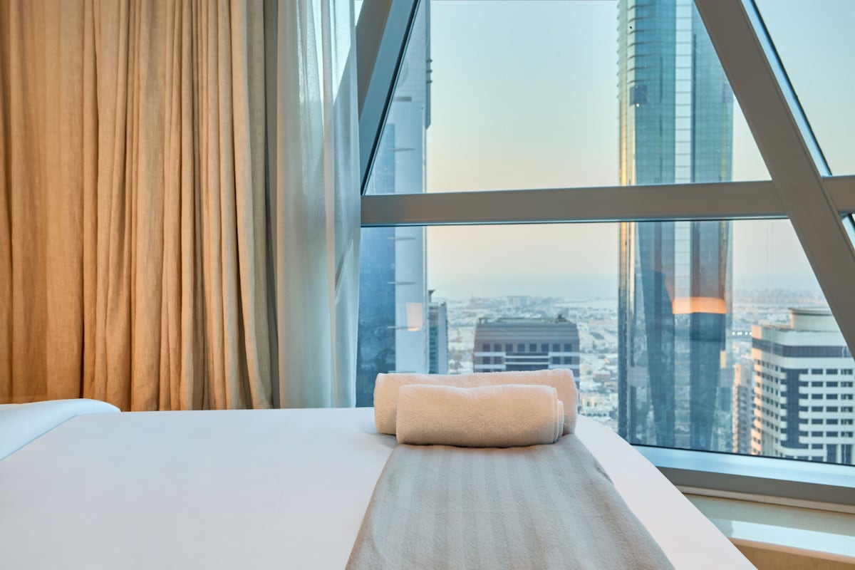 Una cama cuidadosamente preparada se encuentra junto a las ventanas del piso al techo, que ofrecen una vista panorámica de la ciudad y dejan entrar la suave luz de la mañana en la habitación.