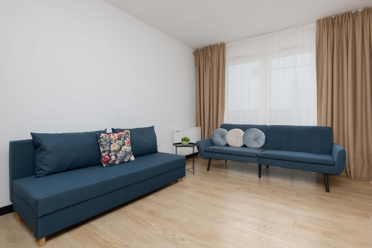 Светла всекидневна с тъмносин диван, разположен срещу бяла стена. Леките дървени подове придават усещане за топлина в помещението. Мека дневна светлина влиза през широк прозорец, покрит с прозрачни завеси.