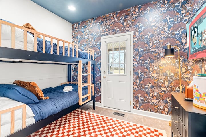 Забавна детска спалня с две двойни кушетки, Roku TV, тапети с елени и игриви килими – уютно място за сън за малки авантюристи, сънуващи под планините.