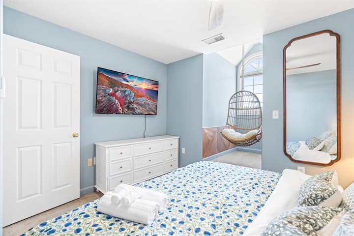 Уютна спалня с голямо двойно легло, смарт телевизор Roku, висящ стол и скрин – очарователно синьо убежище с достатъчно естествена светлина за спокойни престои.