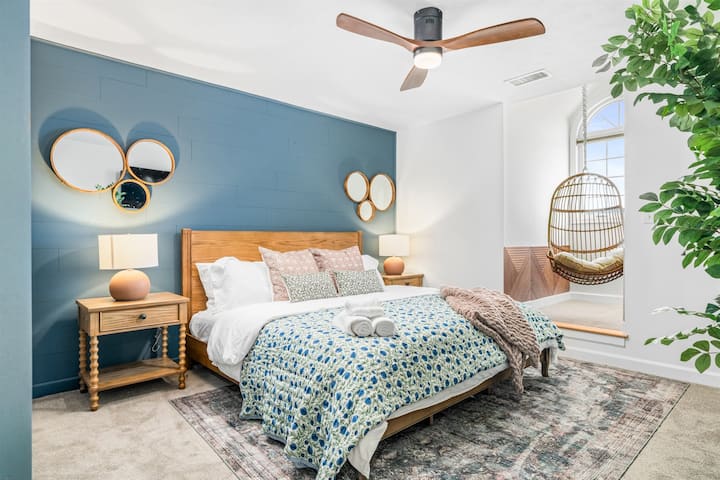 Спалня с голямо двойно легло, вентилатор на тавана, висящ стол и баня – спокойно синьо убежище с естествена светлина и гледка към планината за пълна релаксация.