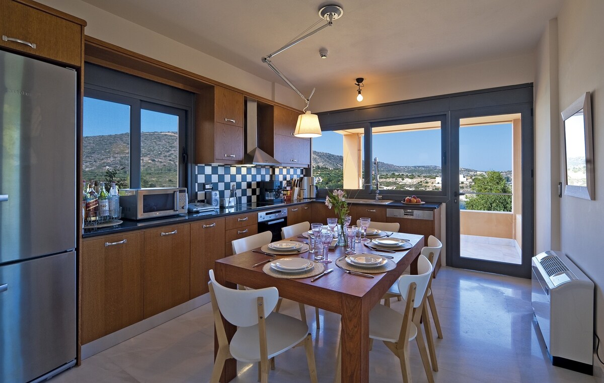 A modern kitchen is presented with wooden cabinetry and a central dining table set for six. The room features large windows with views of the surrounding landscape, and stainless steel appliances including a refrigerator and microwave are visible.