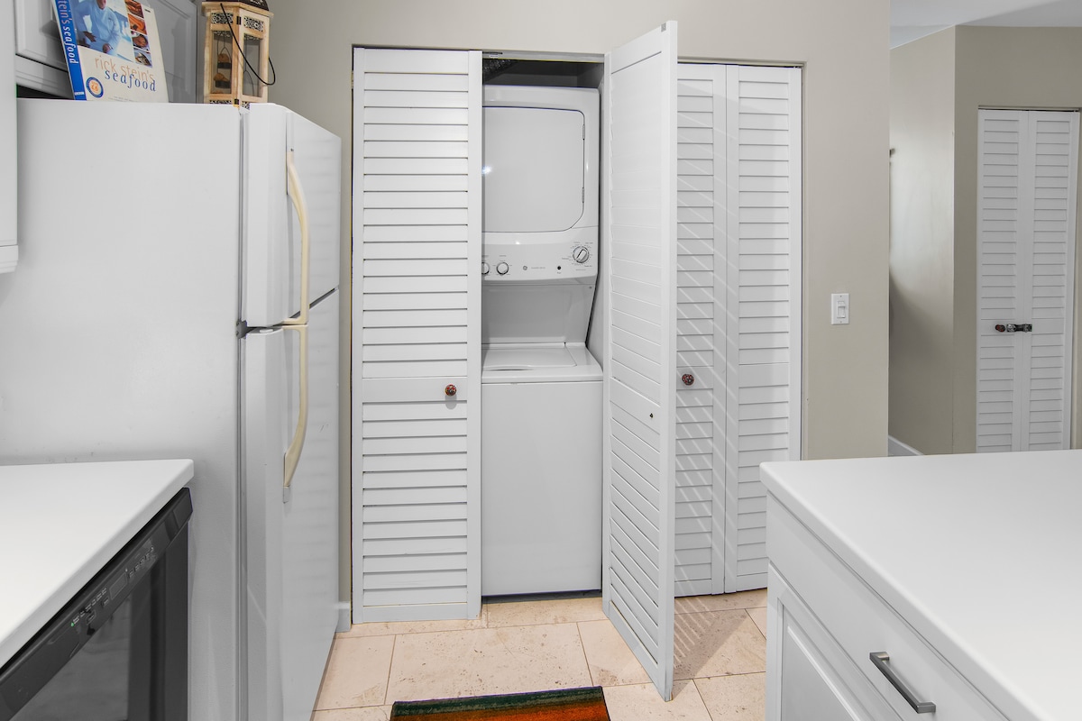 A compact laundry area is neatly integrated into the space, featuring a stacked washer and dryer behind louvered doors. The surrounding kitchen equipment, including a refrigerator and cabinetry, is visible, creating a functional laundry nook.