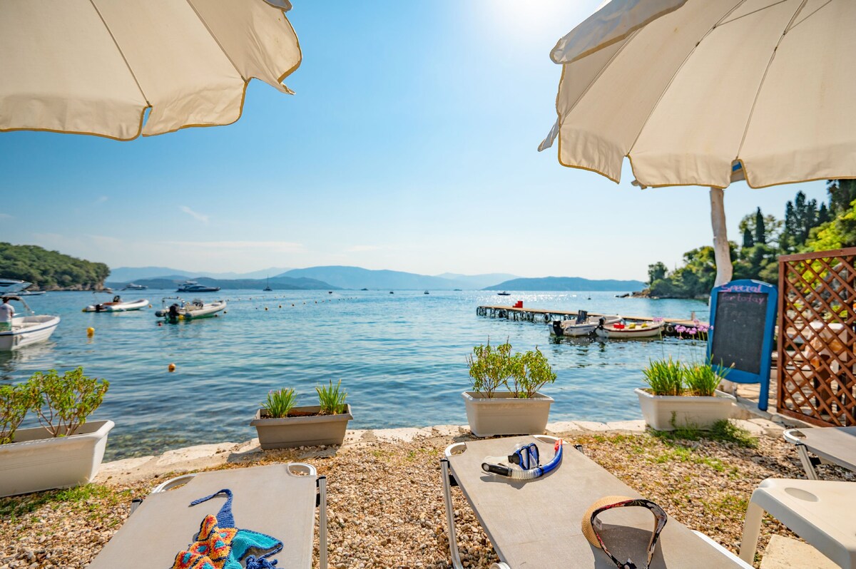 Sunbeds on a pebbly beach are shaded by large umbrellas, offering a view of the tranquil water of Agni Bay. Several boats are anchored nearby, and gentle hills can be seen in the background under a clear blue sky.